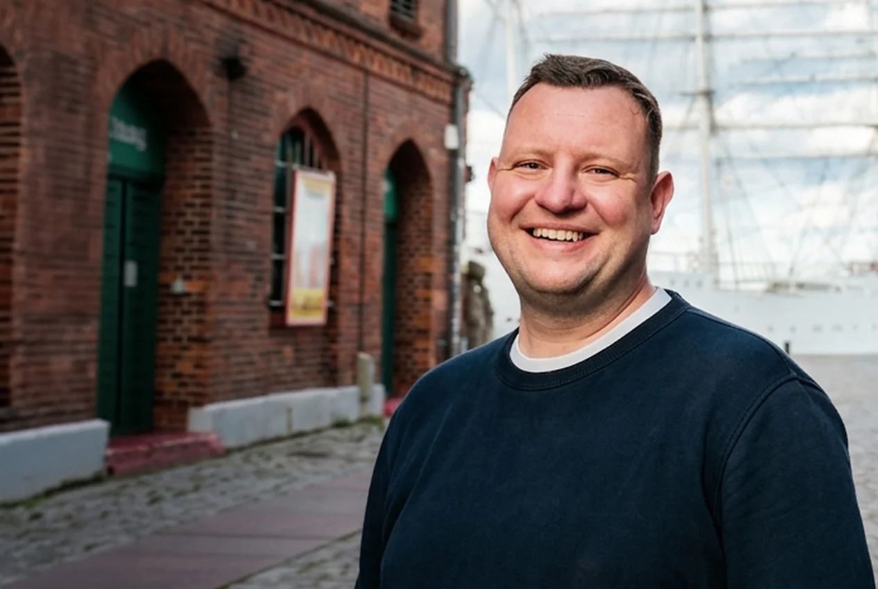 Mathias, Recruiting-Berater bei Talentküste, lächelt in einer Gasse in Stralsund vor einem Segelschiff.