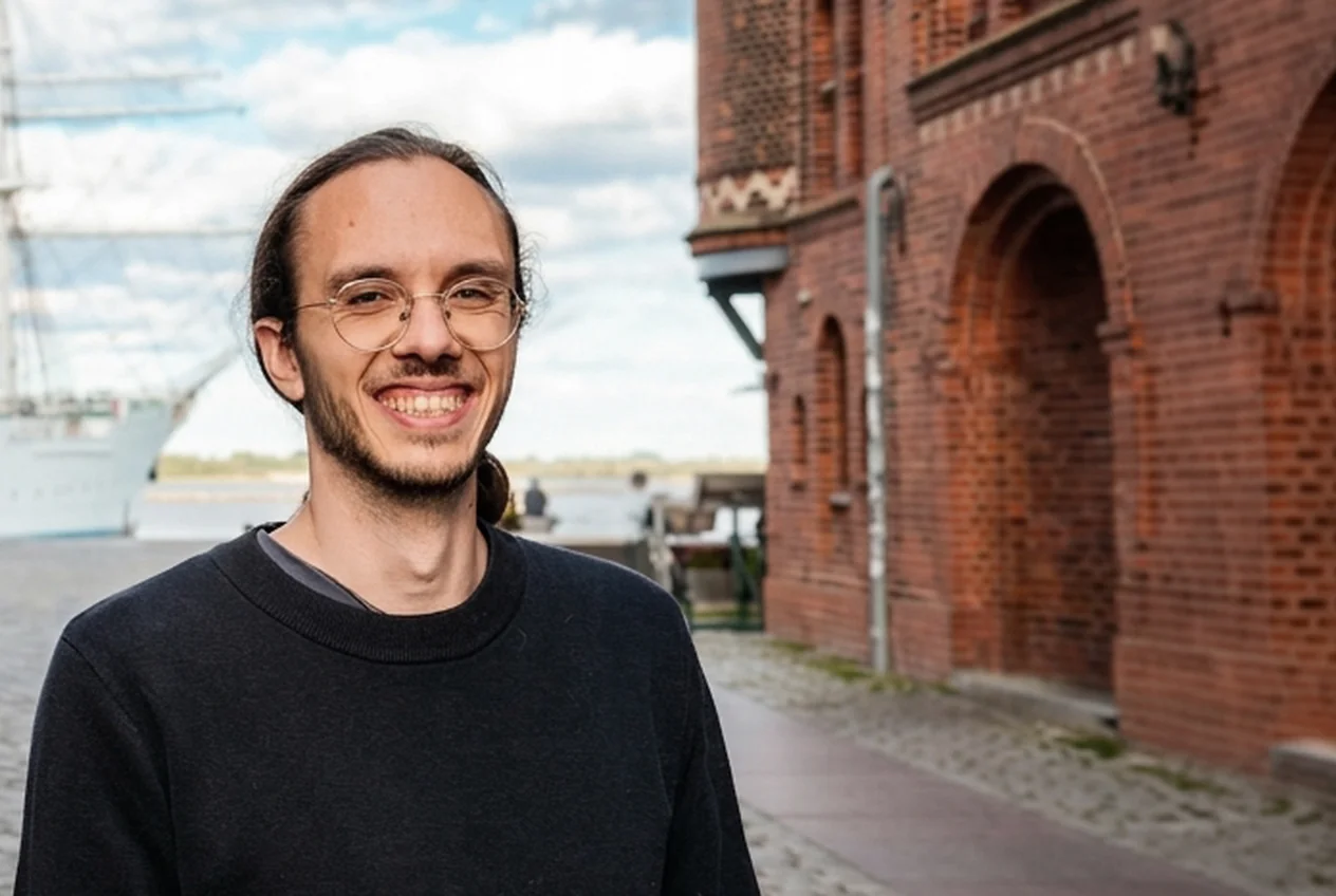 Tom, Marketing-Berater bei Talentküste, lächelt in einer Gasse in Stralsund vor einem Segelschiff.